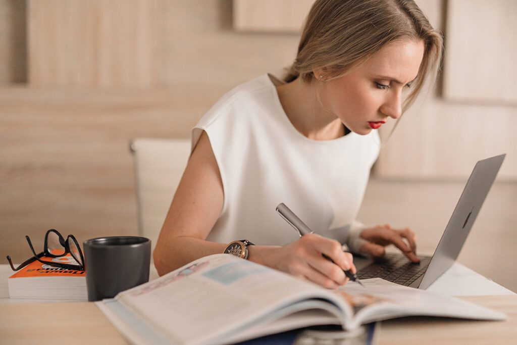 A blonde woman sits at a desk with a notebook and computer while researching branding for her short-term rental.